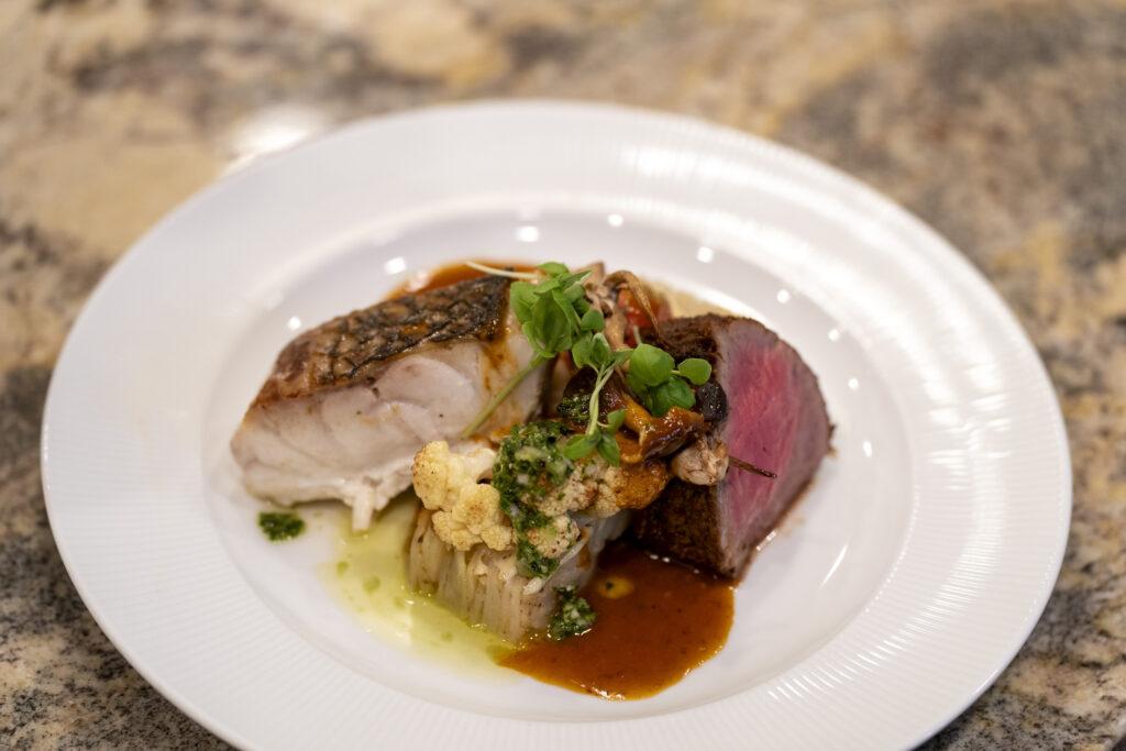 Carved Sous Vide Beef Tenderloin and Stripped Sea Bass, complemented by smoked bacon butter emulsion, roasted cauliflower & chimichurri, herb-scented potato pave, and Cacio e Pepe fondue.